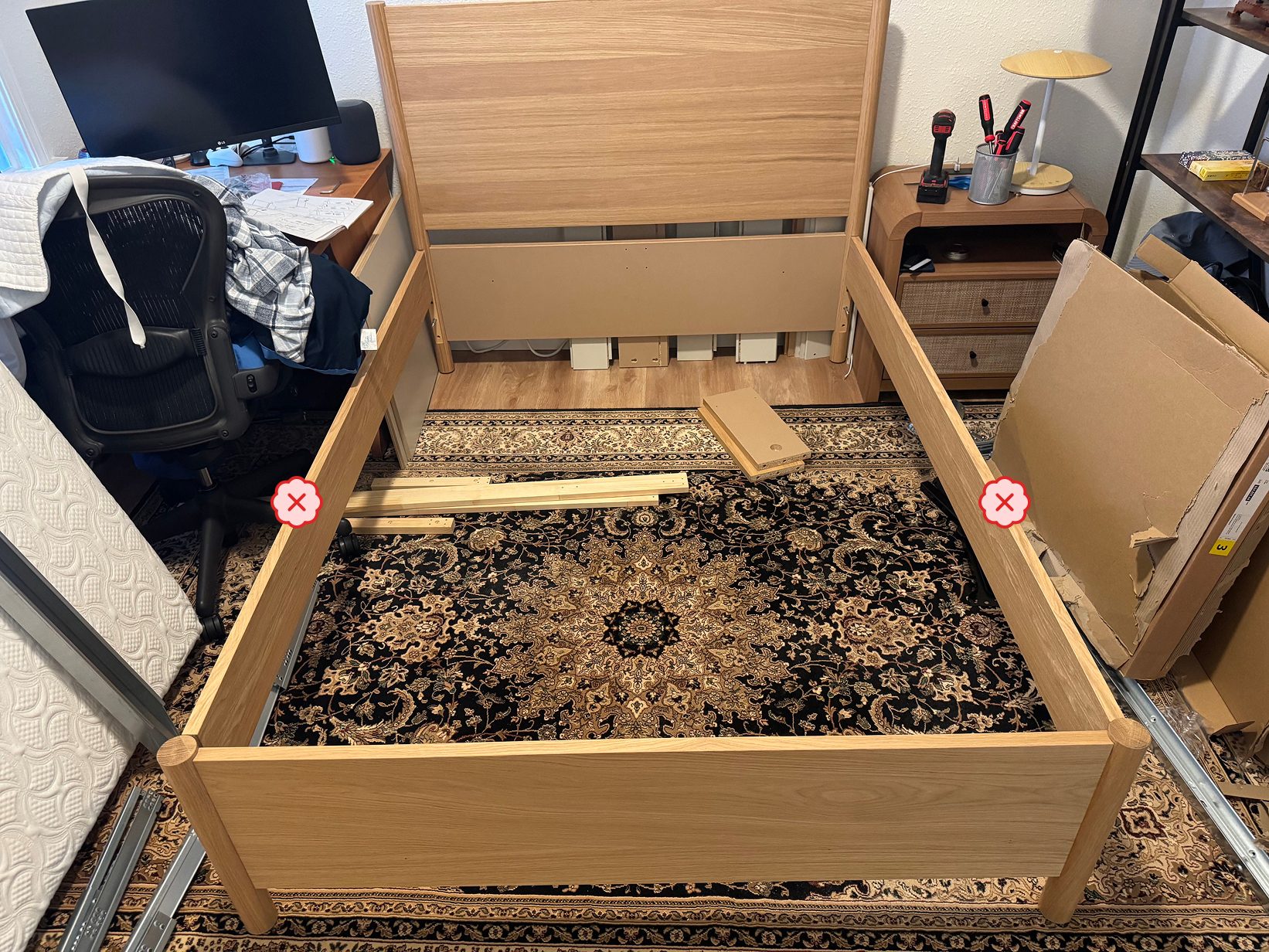 Photograph of a partially assembled wooden bed frame in a bedroom, surrounded by loose slats, torn cardboard packaging, tools, and a mattress leaning nearby, with a red cross on each side rail marking the mistake.