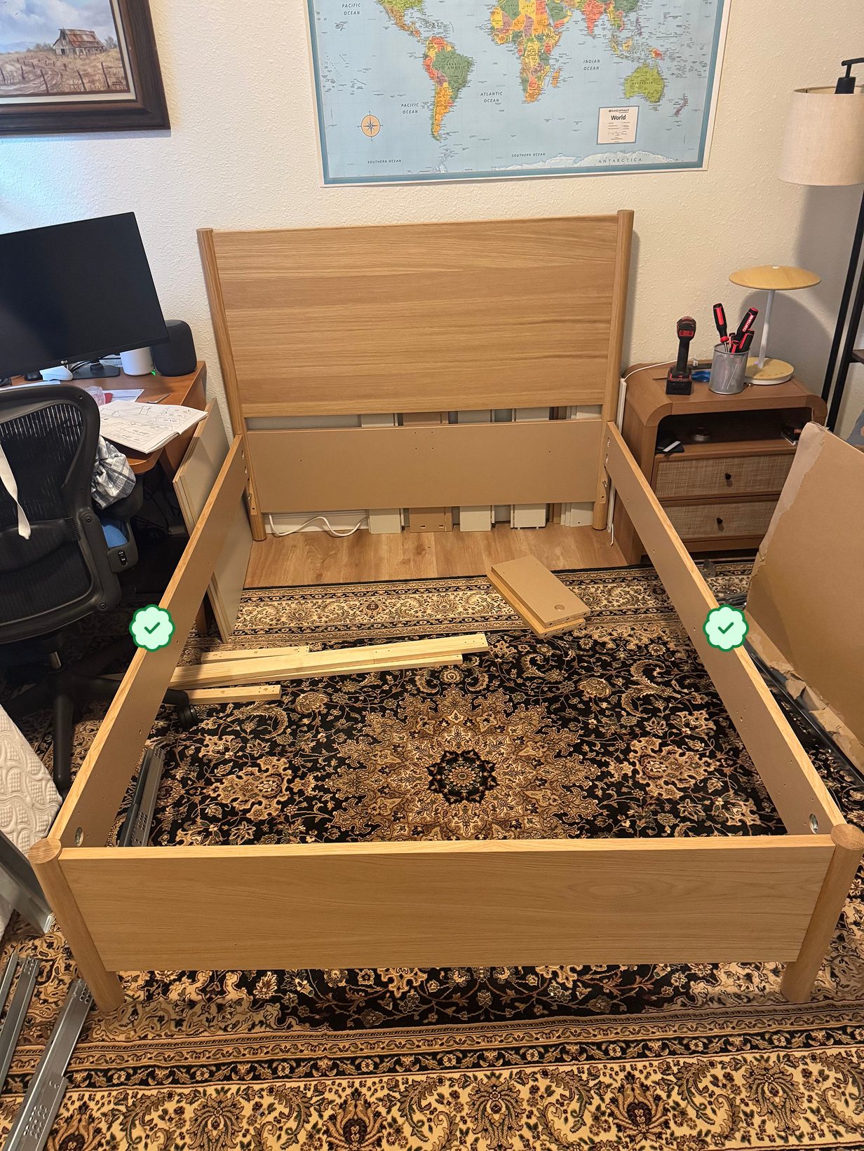 A partially assembled wooden bed frame standing on a patterned rug in a bedroom, with instruction sheets, loose slats, and a power drill nearby, with green checks on both side rails marking them as correctly oriented.
